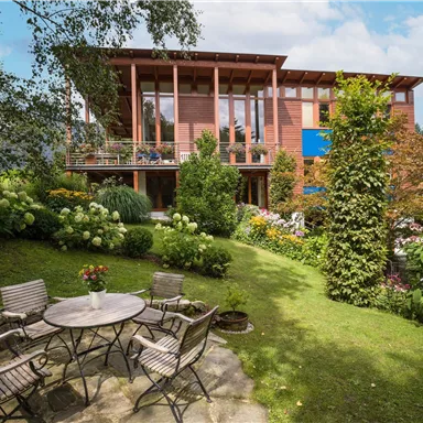 Ein modernes Haus mit großen Fenstern und einem gepflegten Garten. Der Außenbereich ist mit einem Tisch und Stühlen ausgestattet, um eine angenehme Atmosphäre zu schaffen.