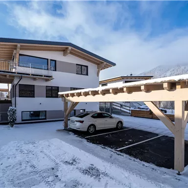 Ein modernes Haus mit einem schneebedeckten Parkplatz und einem überdachten Stellplatz. Im Hintergrund sind Berge und ein blauer Himmel zu sehen.