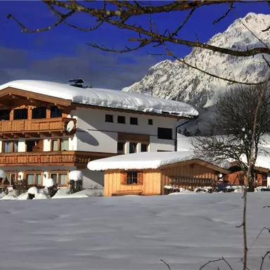 Ein gemütliches Holzhaus im Schnee, umgeben von Bergen. Der Himmel ist klar und der Schnee glänzt in der Sonne.