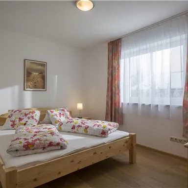 Ein gemütliches Schlafzimmer mit einem Doppelbett und bunten Kissen. Helle Wände und ein großer Fenster sorgen für eine einladende Atmosphäre.