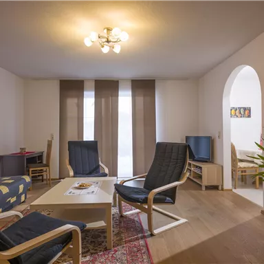 Ein gemütliches Wohnzimmer mit einer bequemen Sitzgruppe und einem Tisch. Große Fenster sorgen für helles Licht und eine entspannte Atmosphäre.