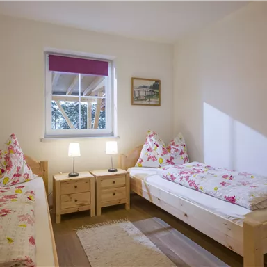 Ein einfaches Schlafzimmer mit zwei Betten und gemütlichen Kissen. Es gibt Nachttische mit Lampen und ein Fenster, das Tageslicht hereinlässt.