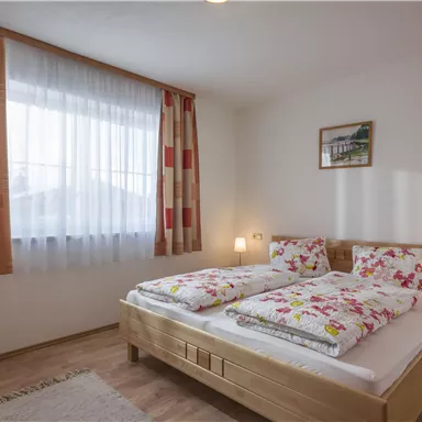 Ein gemütliches Schlafzimmer mit einem Doppelbett und bunten Bettbezügen. Der Raum hat große Fenster mit Vorhängen und ist hell und einladend.