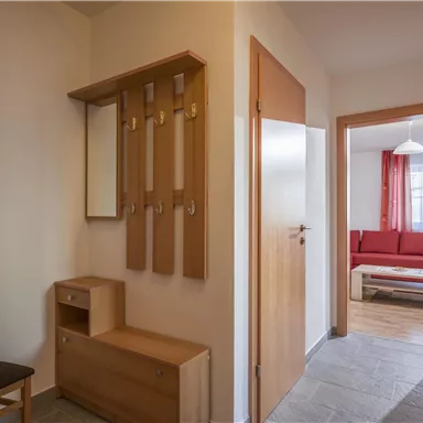 Ein heller Flur mit einer Holzbank und einem Spiegel. Im Hintergrund ist ein Wohnbereich mit rotem Sofa zu sehen.