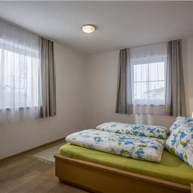 Ein helles Schlafzimmer mit einem Doppelbett und bunten Bettwaren. Große Fenster lassen viel Tageslicht herein.