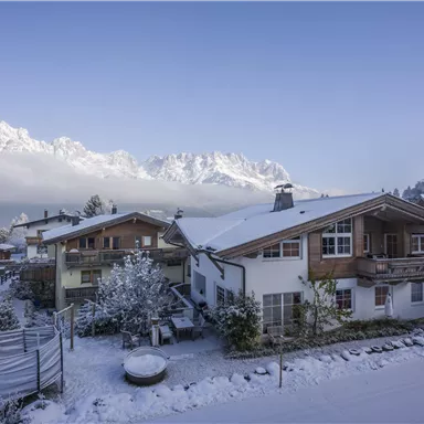 Eine verschneite Landschaft mit mehreren Häusern. Im Hintergrund sind majestätische Berge und ein klarer Himmel zu sehen.