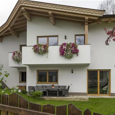 Ein modernes Haus mit blühenden Fensterkästen und einem schönen Garten. Die Fassade ist hell und einladend, umgeben von Bäumen und einer Holzzaun.