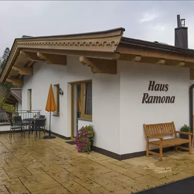 Ein gemütliches Haus mit dem Namen "Haus Ramona". Die Terrasse ist mit Blumen geschmückt und hat einen Tisch mit Stühlen.