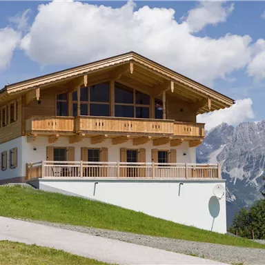 A modern wooden house with large windows and a balcony. In the background, majestic mountains and a blue sky are visible.
