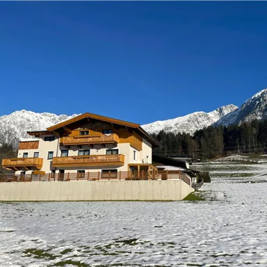 A picturesque house in the mountains, surrounded by snow and a clear blue sky. Snow-capped peaks are visible in the background.