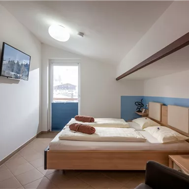 Ein helles Zimmer mit zwei Betten, einem Fernseher und einem Blick aus dem Fenster. Die Einrichtung ist modern und schlicht, mit Pflanzen und dekorativen Elementen.