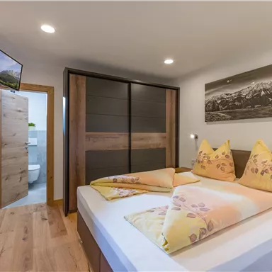 Ein gemütliches Schlafzimmer mit einem Doppelbett und bunten Kissen. Im Hintergrund befindet sich ein geschlossener Schrank und ein Zugang zum Badezimmer.