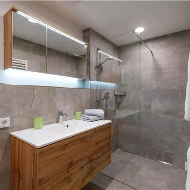 Ein modernes Badezimmer mit einer Glasdusche und einem Holzmöbel. Es gibt eine ansprechende Beleuchtung und frische Handtücher.