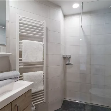 Ein modernes Badezimmer mit einer Dusche und einem Waschbecken. Weißer Fliesenboden und ein Handtuchhalter sind ebenfalls zu sehen.