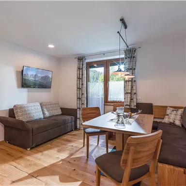 Ein modernes Wohnzimmer mit einem Esstisch und gemütlicher Sitzgelegenheit. Große Fenster sorgen für viel Licht und einen Blick nach draußen.