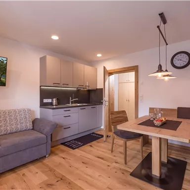 Eine moderne Wohnküche mit einem gemütlichen Sofa und einem Esstisch. Die Einrichtung ist hell und einladend, mit einem Fernseher und einem Uhrwerk an der Wand.