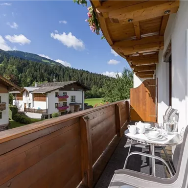 Ein gemütlicher Balkon mit Stühlen und einer Kaffeetasse, umgeben von grünem Bergland. Im Hintergrund sind traditionelle alpine Häuser und ein klarer blauer Himmel zu sehen.