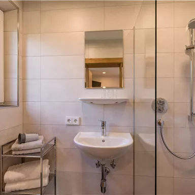 Ein modernes Badezimmer mit einer Dusche und einem Waschbecken. Handtücher liegen ordentlich auf einem Regal.