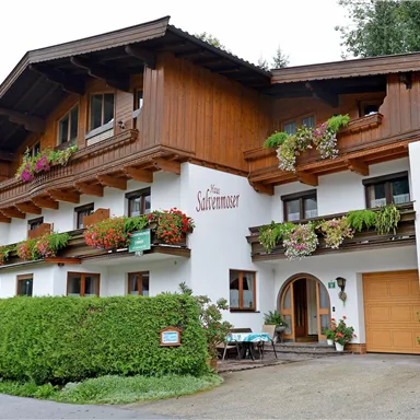 Ein charmantes Haus im alpinen Stil mit Holzverkleidung und bunten Blumen. Es hat mehrere Fenster und einen kleinen Gartenbereich.