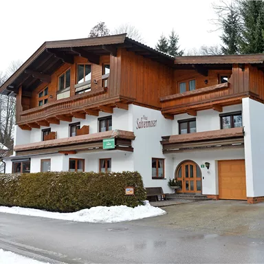 Ein charmantes alpine Gebäude mit Holzverkleidung und großen Fenstern. Es liegt in einer schneebedeckten Umgebung, umgeben von Bäumen.