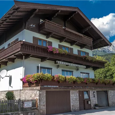 Ein charmantes Berghaus mit mehreren Balkonen und blühenden Pflanzen. Die beeindruckende Landschaft mit Bergen im Hintergrund vervollständigt das Bild.