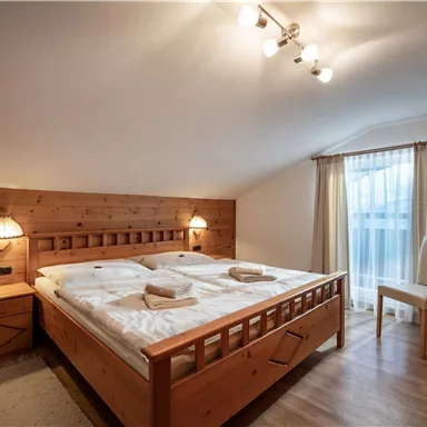 Ein gemütliches Schlafzimmer mit einem Doppelbett aus Holz und warmen Lichtquellen. Fenster lassen viel Tageslicht herein und bieten einen Blick nach draußen.