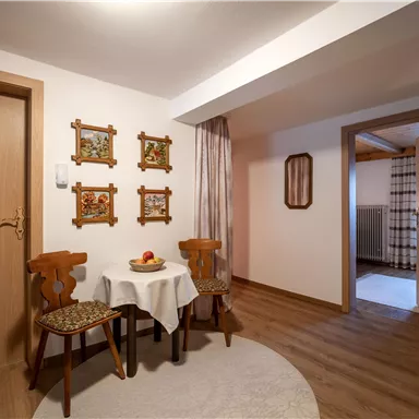 Ein gemütlicher Raum mit einem kleinen Tisch und zwei Stühlen. An der Wand hängen dekorative Bilder, und eine offene Tür führt in einen weiteren Raum.