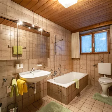 Ein helles Badezimmer mit weißen Fliesen und Holzdecke. Es ist ausgestattet mit einer Badewanne, einem Waschbecken und einem WC, sowie bunten Handtüchern in Gelb und Grün.