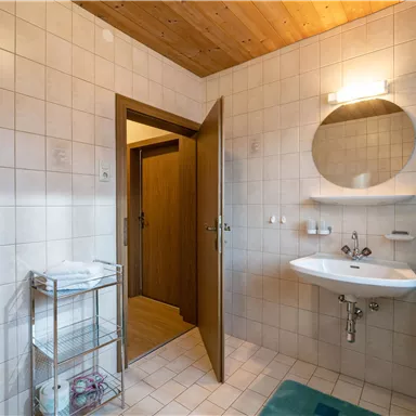 Ein modernes Badezimmer mit hellen Fliesen und Holzdecke. Es gibt ein Waschbecken, einen Spiegel und einen offenen Türbereich.