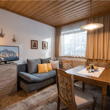 Ein gemütliches Wohnzimmer mit einem Fernseher, einer Sitzecke und einem Esstisch. Die Wände sind hell, und die Fenster lassen viel Licht herein.