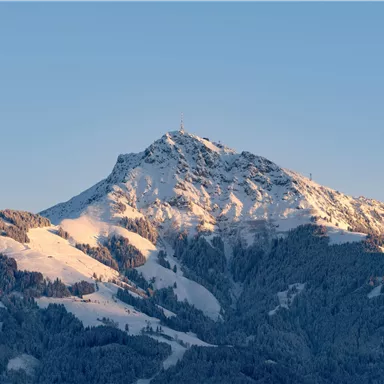 Ein majestätischer Berg mit schneebedecktem Gipfel und sanften Hängen. Der Himmel ist klar und strahlt in sanften Farbtönen.