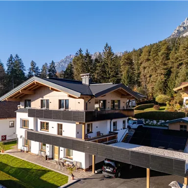 A modern building surrounded by trees and mountains. The architecture is inviting with a clear blue sky in the background.