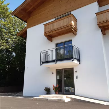 Ein modernes Haus mit Holzverkleidung und einem Balkon. Vor der Eingangstür stehen Blumen in einem Topf.
