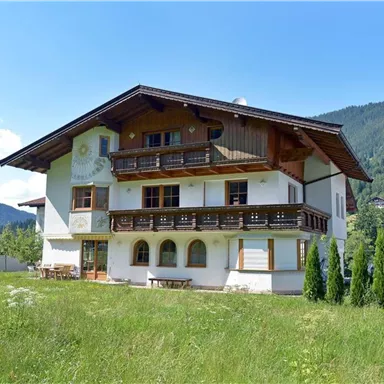 A charming alpine house with balconies and a green garden. In the background, gentle hills and a blue sky can be seen.