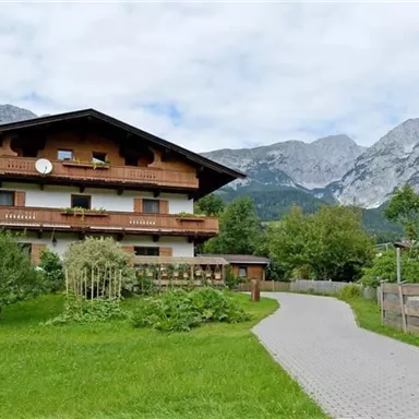 Ein traditionelles Holzhaus umgeben von grüner Landschaft und Bergen. Der Himmel ist bewölkt, und ein Weg führt zur Haustür.