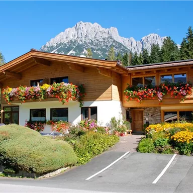 A beautiful, traditional wooden house with colorful flowers in front of the facade. In the background, an impressive mountain rises under a clear blue sky.