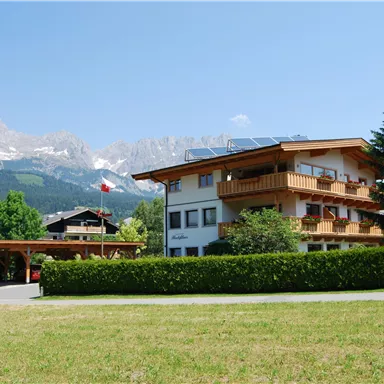 Ein charmantes Gebäude mit Holzverkleidung und Balkonen in einer grünen Umgebung. Im Hintergrund erheben sich majestätische Berge unter klarem Himmel.