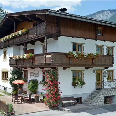 Ein traditionelles Haus mit einem Holzbalkon und vielen Blumen. Im Vordergrund befindet sich eine Terrasse mit einem Sonnenschirm.