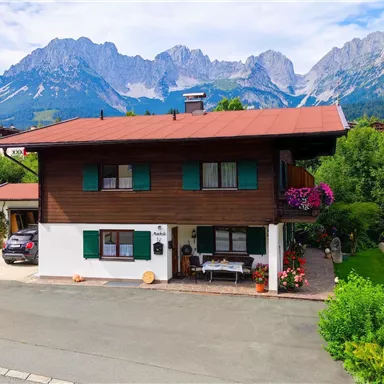 Ein wunderschönes, rustikales Holzhaus vor majestätischen Bergen. Der Garten ist grün und blühend, ideal für entspannte Momente.