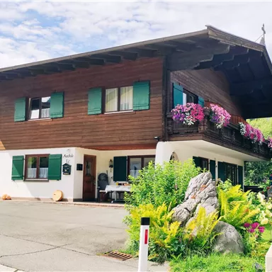 Ein traditionelles Holzhaus mit grünen Fensterläden und Blumenkästen. Der Garten ist üppig bewachsen und die Umgebung wirkt malerisch.