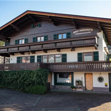 Ein traditionelles alpenländisches Haus mit Holzelementen und grünen Fensterläden. Die Fassade zeigt einen einladenden Balkon und ist von einer gepflegten Hecke umgeben.