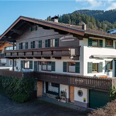 Ein traditionelles Ferienhaus in den Alpen mit einem großen Balkon und vielen Fenstern. Der Garten ist grün und gepflegt, mit Blick auf die umliegenden Berge.