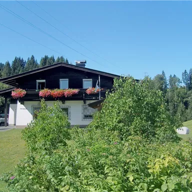 Ein charmantes Bauernhaus umgeben von üppigem Grün und blühenden Pflanzen. Im Hintergrund sind Wälder und ein klarer blauer Himmel zu sehen.