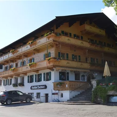 Ein traditionelles Gebäude im alpinen Stil mit mehreren Balkonen und bunten Blumen. Es befindet sich in einer ruhigen Umgebung mit grünen Bäumen und einer klaren blauen Himmel.
