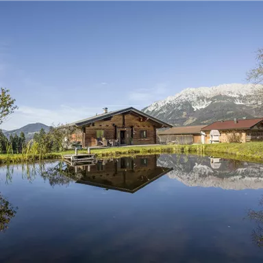 Ein malerisches Holzhaus am Rand eines klaren Teichs. Im Hintergrund sind beeindruckende Berge unter einem klaren Himmel zu sehen.