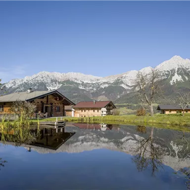 Eine malerische Landschaft mit einem ruhigen Teich und einem Blick auf schneebedeckte Berge. Im Vordergrund steht ein traditionelles Holzhaus.