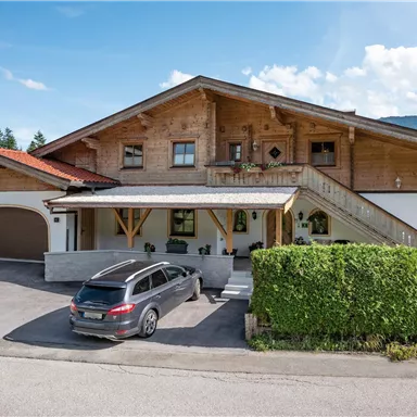 Ein großes Holzhaus mit einem Dach und einer breiten Veranda. Vor dem Haus steht ein grauer Kombiwagen und es gibt viel Grün im Hintergrund.