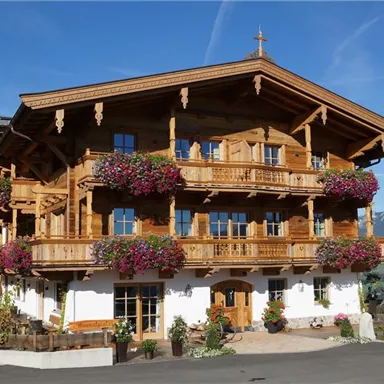 A traditional wooden chalet with many flowers on the balconies. In the background, mountains and a clear sky are visible.