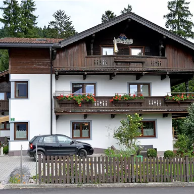 A two-story house in alpine style with a balcony and blooming windows. In the foreground, there is a car, and there is a small garden with a fence.