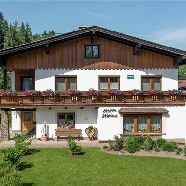 A cozy house in alpine style with wooden cladding and flower boxes. Surrounded by green forests and an inviting meadow.